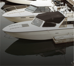Photograph of several white boats docked in dark water. The closest boat has a black canvas cover over its cabin, and its reflection is visible in the water. A wooden dock is on the right.