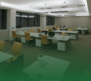 Photograph of a modern, empty open-plan office with rows of light-colored desks, each equipped with a computer monitor and a white desk phone. Orange ergonomic office chairs are pulled up to each workstation. The room has brown carpet, bright overhead lighting, and large windows along the back wall showing darkness outside.