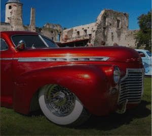 Photograph of a shiny, deep red vintage car with chrome trim, wire wheels, and whitewall tires, parked on green grass in front of the stone ruins of an old castle under a blue sky.