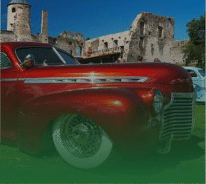 Photograph of a shiny, deep red vintage car with chrome trim, wire wheels, and whitewall tires, parked on green grass in front of the stone ruins of an old castle under a blue sky.