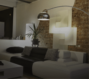 Photograph of a modern living room with a two-tone sectional sofa (dark brown and white), a white coffee table, and an arched chrome floor lamp. Behind the sofa, a potted plant sits on a shelf, and geometric white wall art is illuminated against a brick wall.