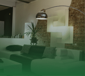 Photograph of a modern living room with a two-tone sectional sofa (dark brown and white), a white coffee table, and an arched chrome floor lamp. Behind the sofa, a potted plant sits on a shelf, and geometric white wall art is illuminated against a brick wall.