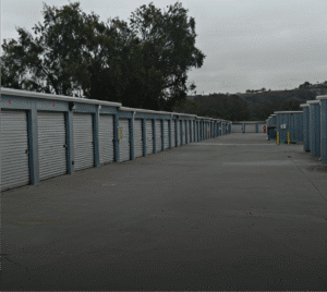 Photograph of a long row of self-storage units with corrugated metal doors and light blue frames, extending into the distance on a concrete driveway. More units are visible in the background, with hills and trees under an overcast sky.