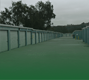 Photograph of a long row of self-storage units with corrugated metal doors and light blue frames, extending into the distance on a concrete driveway. More units are visible in the background, with hills and trees under an overcast sky.