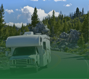 Photograph of a white RV driving on a winding road through a forest with large rocks and a blue sky with white clouds.