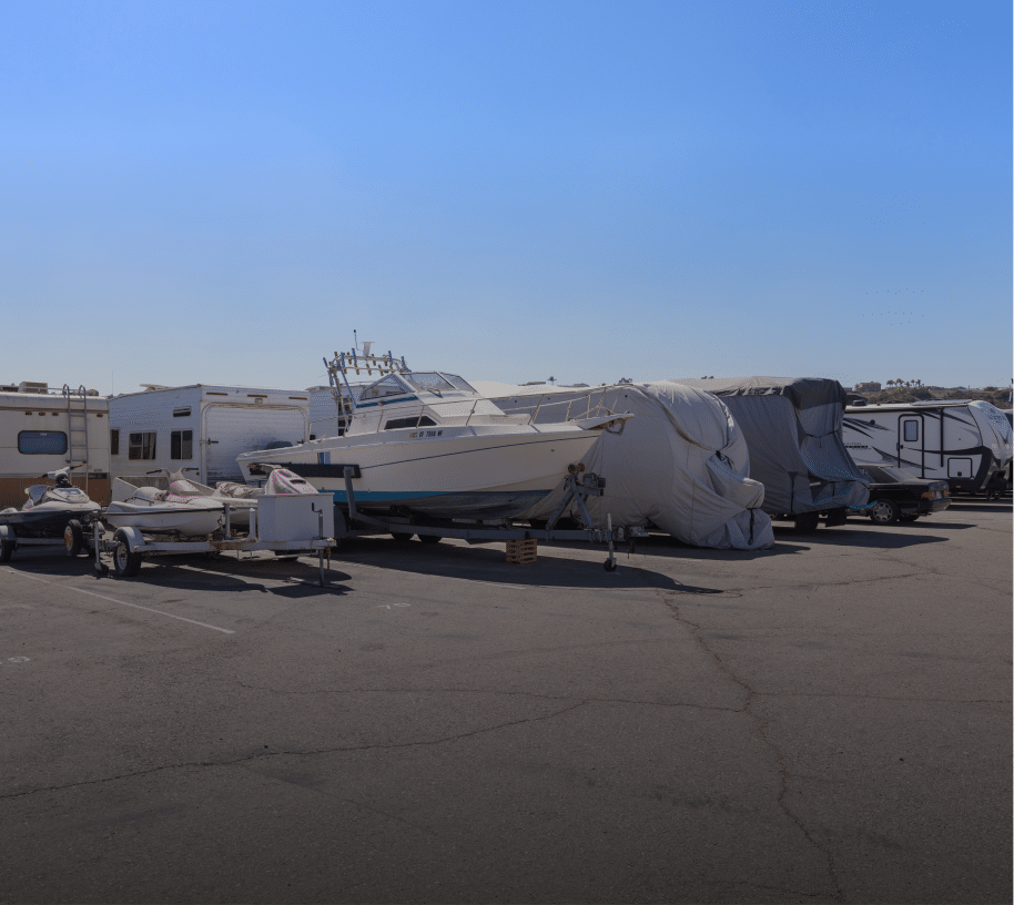 Outdoor storage lot with various recreational vehicles including boats, jet skis, and covered RVs parked on asphalt under a clear blue sky, showcasing secure vehicle and marine storage facility.