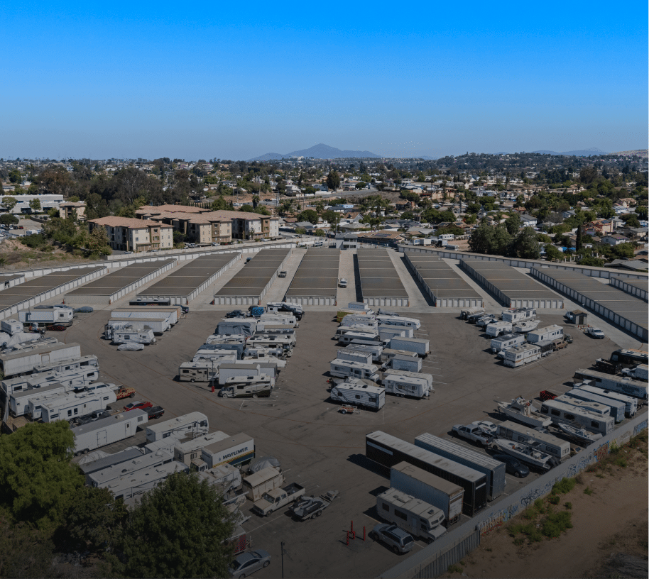 Aerial view of a large RV and boat storage facility featuring multiple rows of covered and uncovered storage units. Numerous recreational vehicles, trailers, and motorhomes are neatly parked in open spaces, with long rows of storage garages visible in the background.
