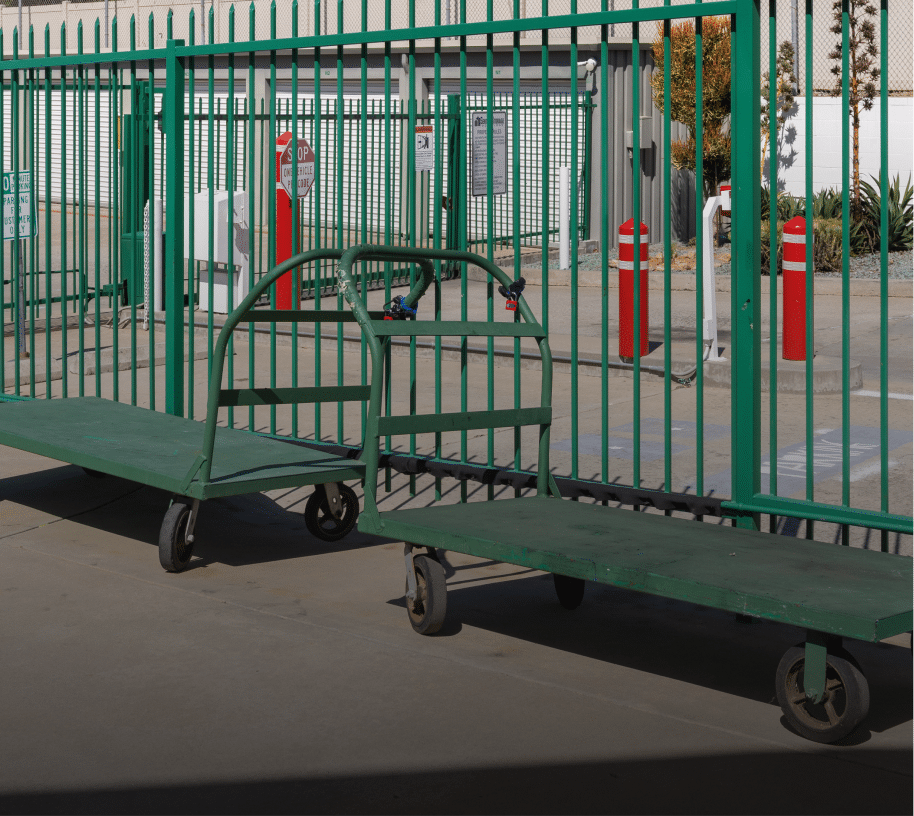 Green metal flatbed carts with wheels positioned beside a tall green security fence at a gated self-storage facility, with red bollards, keypad entry system, and roll-up storage unit doors.