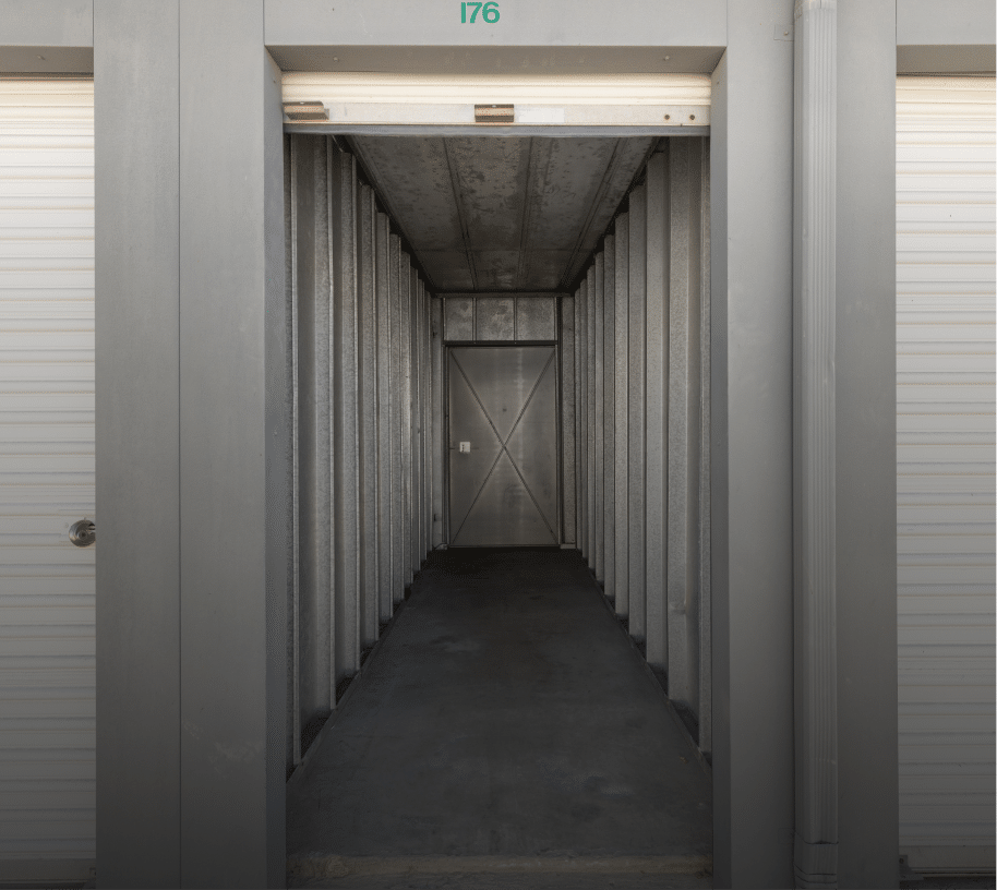 Interior view of an empty metal self-storage unit with corrugated steel walls, concrete floor, and roll-up door. The space is clean, narrow, and well-lit, designed for secure storage of personal or business belongings.