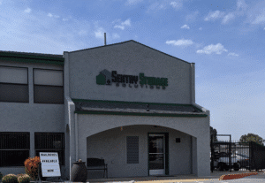 Photograph of the exterior of a Sentry Storage Solutions building. The grey stucco building has a prominent sign on its gable that reads "Sentry Storage Solutions" with a house and person icon. Below, an arched entrance leads to the main door.