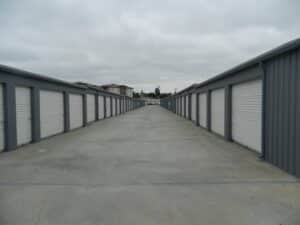 Photograph of a long, straight concrete aisle between two rows of self-storage units. The units have gray frames and white corrugated roll-up doors. The path recedes into the distance under a cloudy sky, with buildings visible in the background.